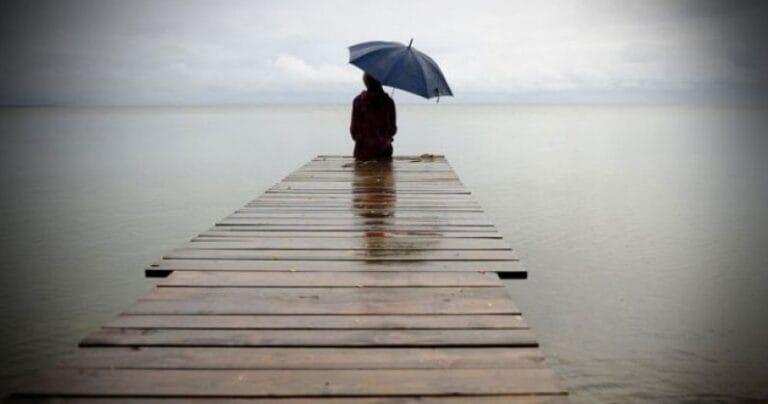 Ilustrasi seorang yang duduk di jembatan di bibir pantai saat hujan turun